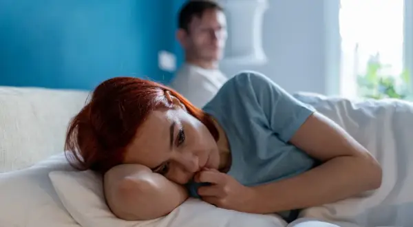 Smutna kobieta leży w łóżku, czując się jak ofiara w związku. Jej partner siedzi za nią, odwrócony plecami.