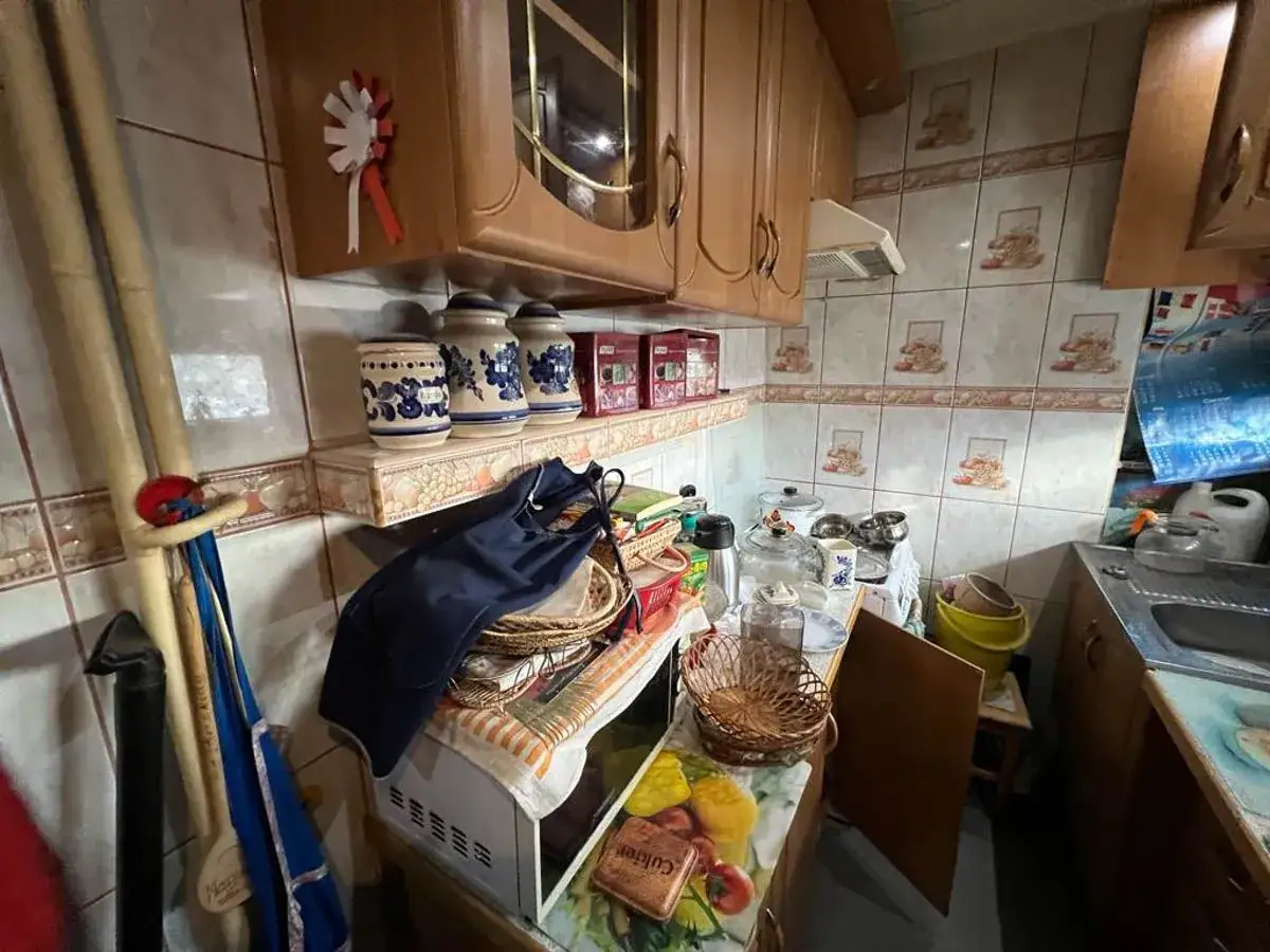 Kuchnia w stanie chorobliwego bałaganiarstwa. Na półkach stoją słoje, fartuch, koszyki, a na blacie piętrzą się naczynia i inne przedmioty.